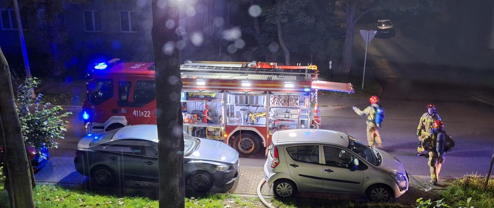 Na zdjęciu widać nocną akcję straży pożarnej na miejskiej ulicy. Na pierwszym planie stoi wóz strażacki z otwartymi bocznymi schowkami, w których widać sprzęt ratowniczy, węże, narzędzia i gaśnice. Obok wozu zaparkowane są samochody osobowe, a w tle widać trzech strażaków w pełnym umundurowaniu i hełmach, z czego jeden ma na sobie aparat powietrzny. Teren akcji jest dobrze oświetlony reflektorem i zabezpieczony taśmą. Panuje noc, a światła pojazdów ratunkowych nadają scenie niebiesko-czerwony blask.