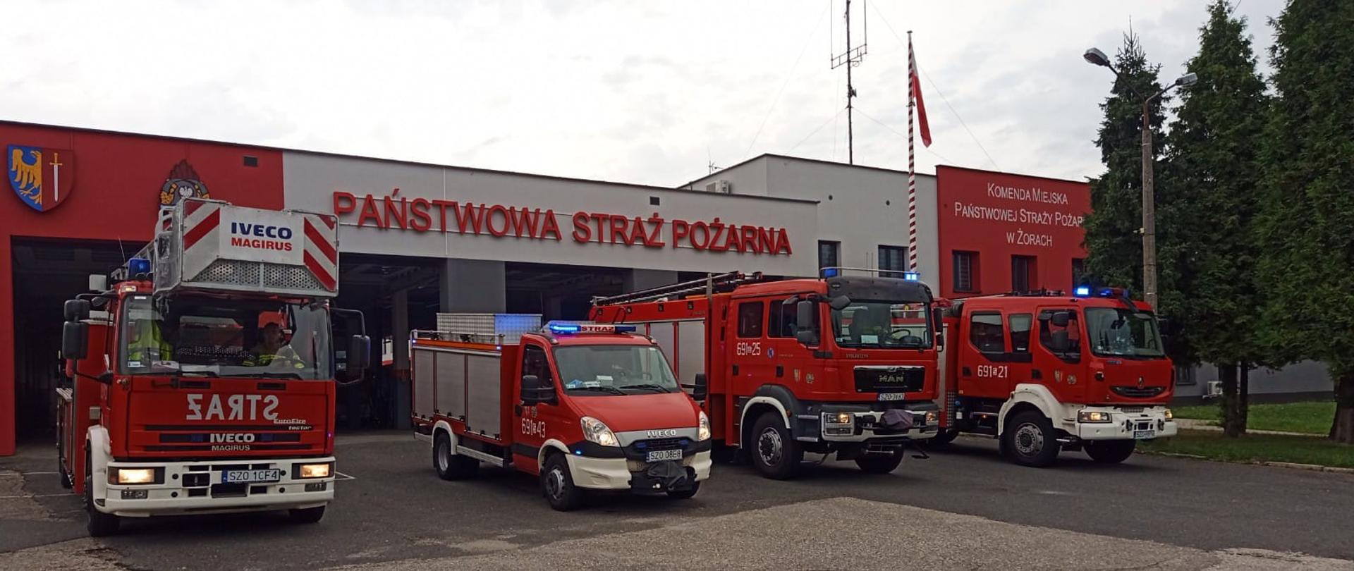 Cztery samochody strażackie ustawione w rzędzie przed garażem Komendy. W tle budynek oraz w prawej części zdjęcia kilka drzew iglastych.
