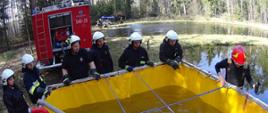 Zdjęcie ukazuje strażaków w trakcie ćwiczeń. Na pierwszym planie widać rozłożony zbiornik na wodę, w którym pływa pompa i pompuje wodę. Dookoła zbiornika strażacy obserwują przebieg ćwiczeń. W tle widać samochód strażacki, a na dalszym planie grupę strażaków przy innych ćwiczeniach. Po prawej stronie zdjęcia zbiornik wodny.