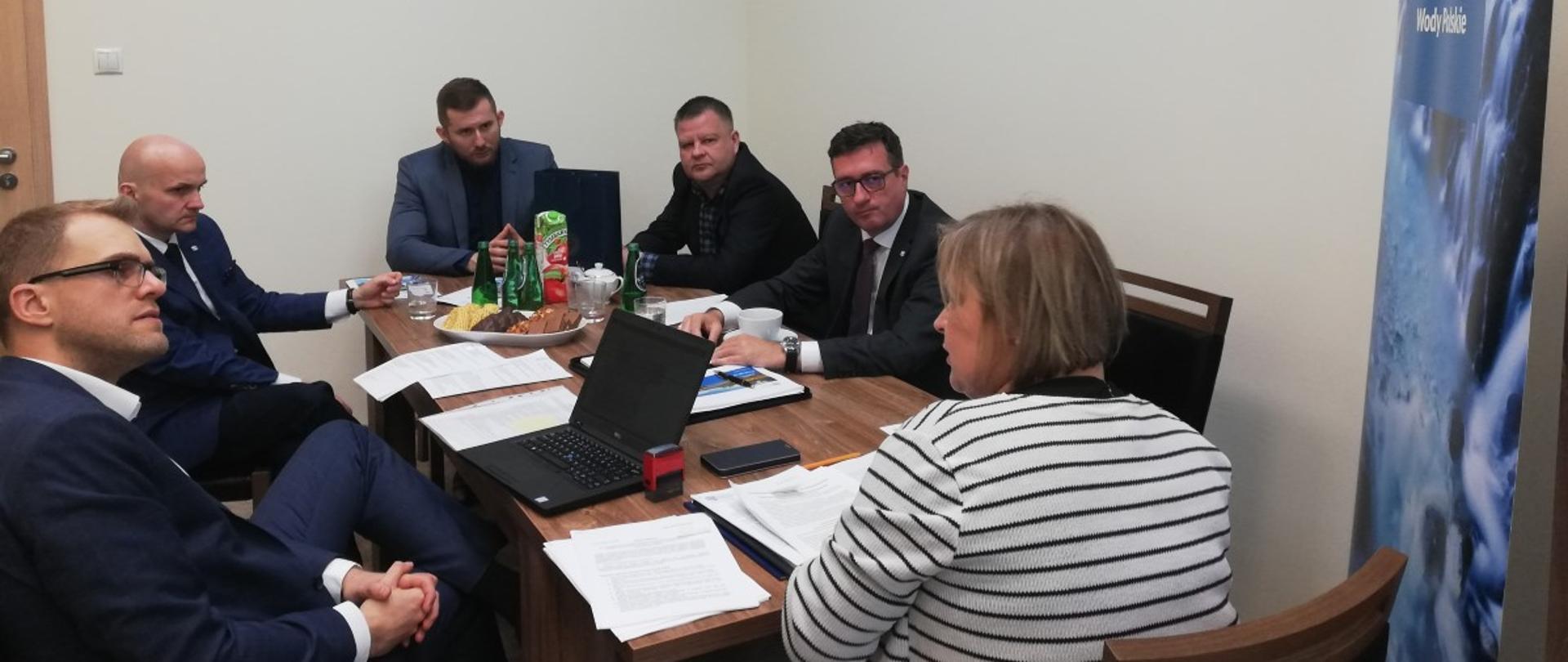 Jasny pokój ze stołem na środku. Przy stole siedzi kilka osób, w tym Pan Przemysław Daca i Pani Agnieszka Szymula.