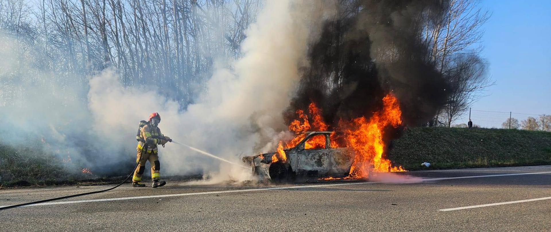 Na drodze płonie auto osobowe, duże płomienie i dużo dymu. Z lewej strony strażak gasi pożar.