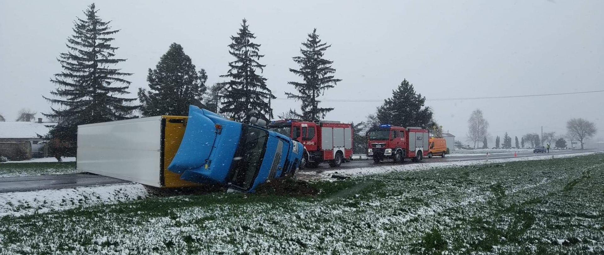 Strażak OSP przechodzący obok leżącej na jezdni na boku naczepy samochodu ciężarowego całkowicie blokującej ruch na drodze, w tyle za naczepą stojący samochód gaśniczy straży pożarnej, widoczny na poboczu drogi śnieg.