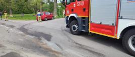 Na zdjęciu na pierwszym planie widoczny jest pojazd pożarniczy. Na drugim planie widać uszkodzony pojazd koloru czerwonego oraz strażaka, który wypełnia dokumentacje. 