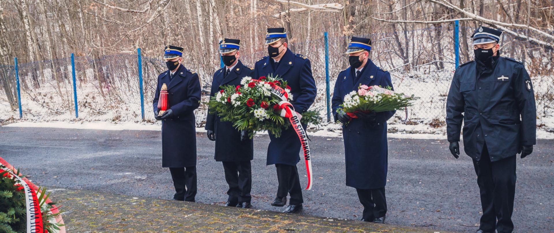 Na zdjęciu widoczni są od lewej bryg. Arkadiusz Korzeniewski - komendant miejski PSP w Katowicach, st. bryg. Mirosław Synowiec - zastępca śląskiego komendanta wojewódzkiego PSP, bryg. Paweł Nalepka - komendant miejskiego PSP w Chorzowie, bryg. Arkadiusz Labocha - komendant miejski PSP w Świętochłowicach oraz ks. Michał Palowski - kapelan strażaków województwa śląskiego. Osoby są ubrane w mundury wyjściowe i maseczki ochronne i składają wieńce i znicz pod pomnikiem upamiętniającym ofiary katastrofy w hali Międzynarodowych Targów Katowickich.