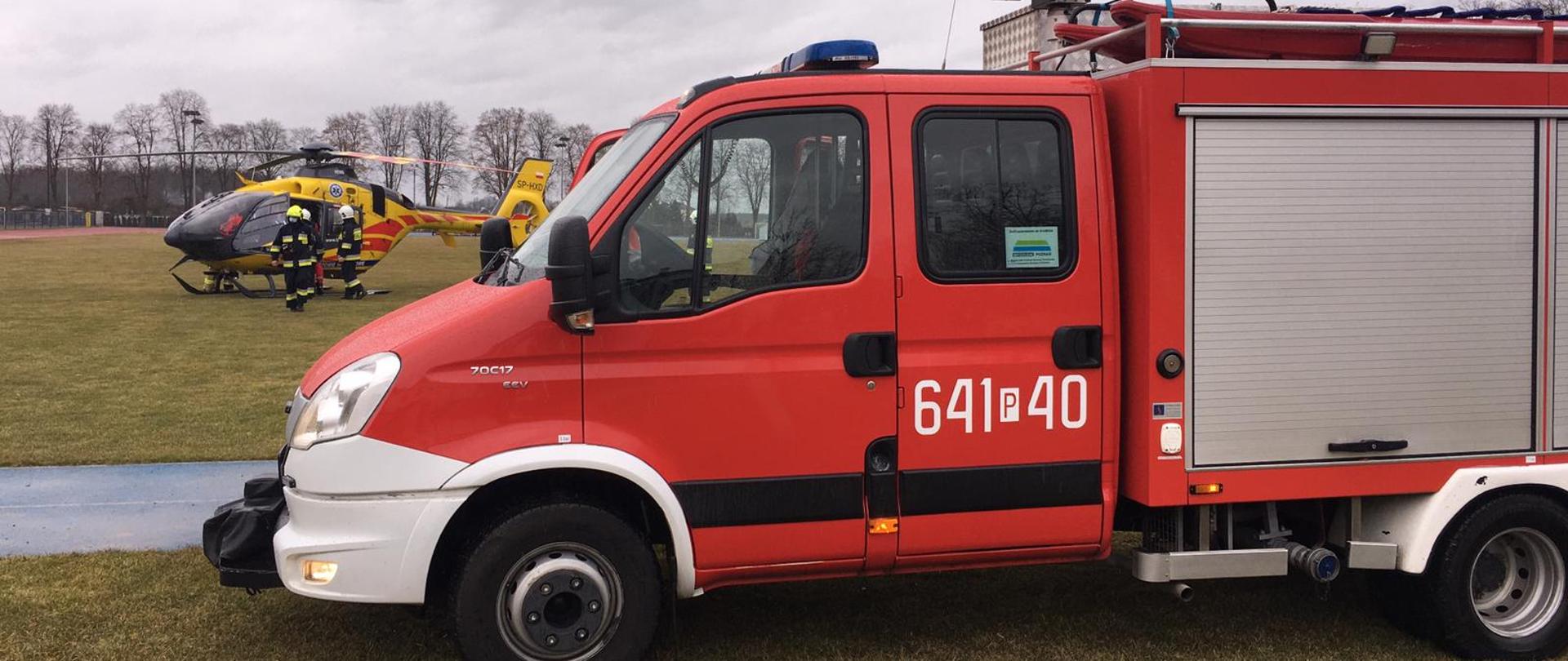 Wóz strażacki na pierwszym planie, w tle śmigłowiec LPR oraz strażacy.