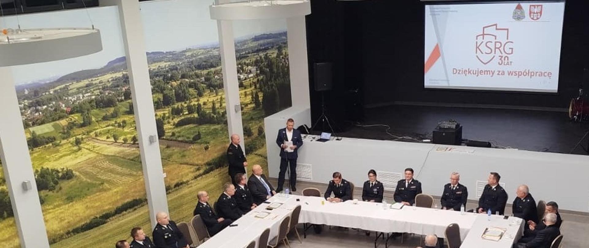 Na zdjęciu widać uroczyste spotkanie strażaków z okazji 30-lecia Krajowego Systemu Ratowniczo-Gaśniczego (KSRG).
Uczestnicy — osoby w umundurowaniu strażackim — siedzą przy stołach ustawionych w kształt dużej litery U. Na stołach leżą dyplomy i dokumenty, które wyglądają na wyróżnienia wręczane podczas wydarzenia. Przed uczestnikami stoją również filiżanki, szklanki i napoje, co sugeruje oficjalne spotkanie połączone z częścią towarzyską.
Z przodu sali, przy scenie, stoi mężczyzna w garniturze przemawiający do zebranych. Za nim, na ekranie, wyświetlona jest grafika z logo KSRG i napisem „30 lat – Dziękujemy za współpracę”.
Ściany sali zdobią duże panoramy krajobrazów, co nadaje przestrzeni jasny i elegancki wygląd.
