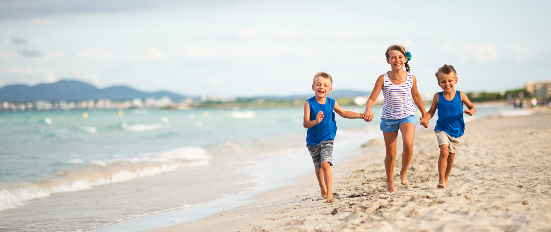 Po plaży w prawym rogu biegnie trójka dzieci dziewczynka i dwóch chłopców trzymających się za ręce