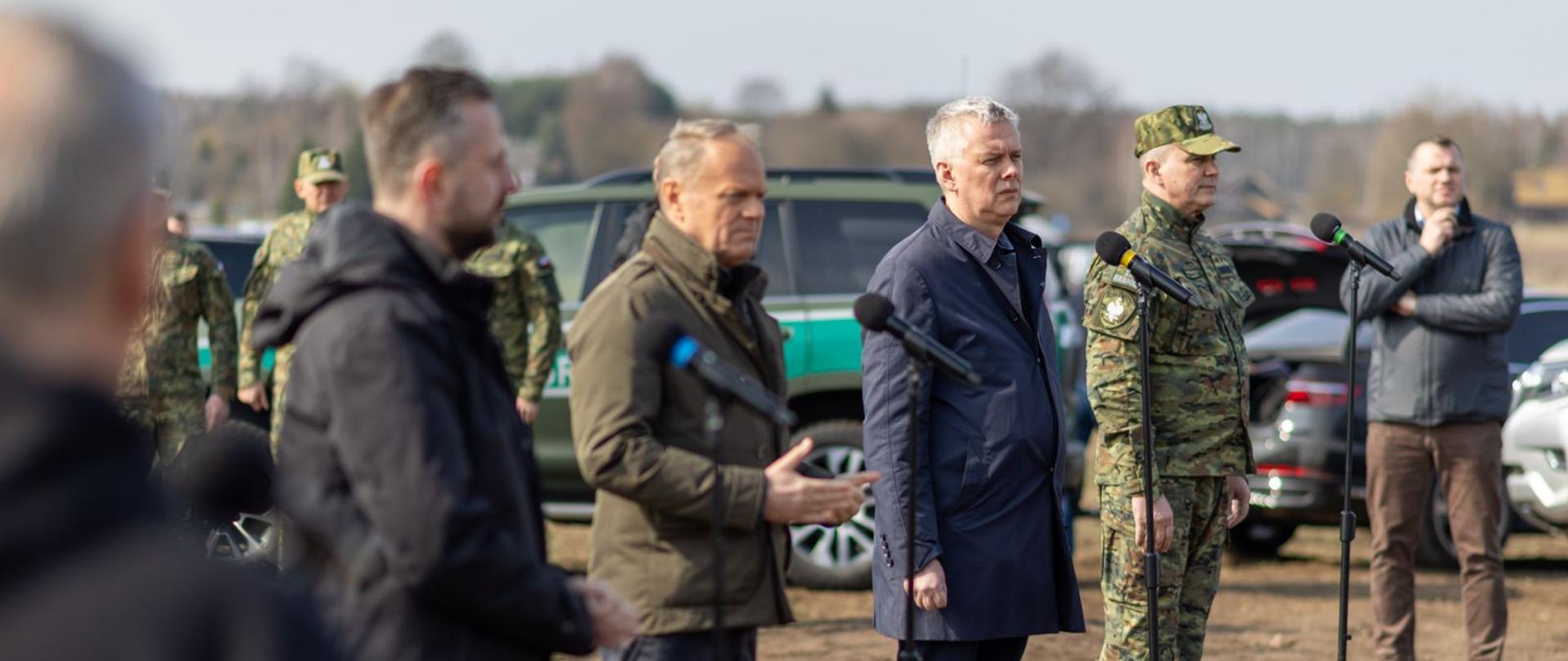 Na zdjęciu od lewej szef MON Władysław Kosiniak-Kamysz, premier Donald Tusk, minister Tomasz Siemoniak, komendant SG Robert Bagan. Uczestnicy konferencji prasowej stoją przed czarnymi mikrofonami. W tle Straż Graniczna i samochody SG. 