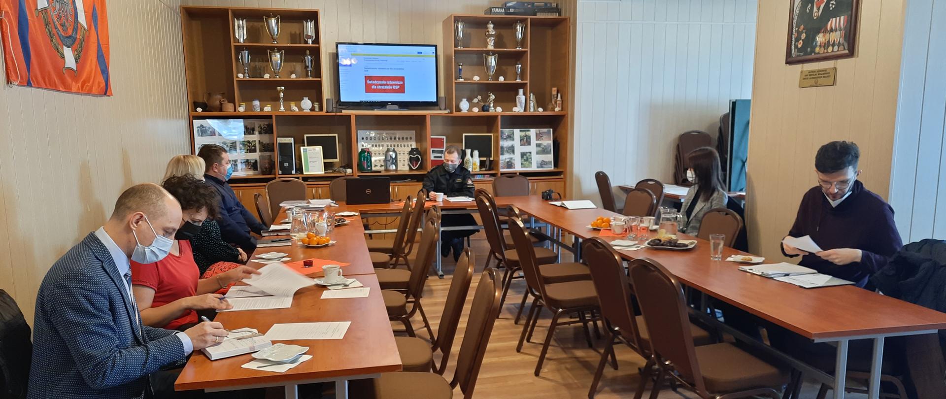 zdjęcie zrobione w świetlicy OSP Sępólno Krajeńskie. Przy stołach ustawionych w literę U zwróconą do patrzącego siedzą po lewej i prawej stronie przedstawiciele samorządów z powiatu sępoleńskiego. Naprzeciwko patrzącego siedzi Komendant Powiatowy PSP, który prowadzi szkolenie. Nad Komendantem znajduje się telewizor na którym widoczna jest strona internetowa Komendy Głównej PSP. Na stołach dokumenty oraz mandarynki i ciastka. 