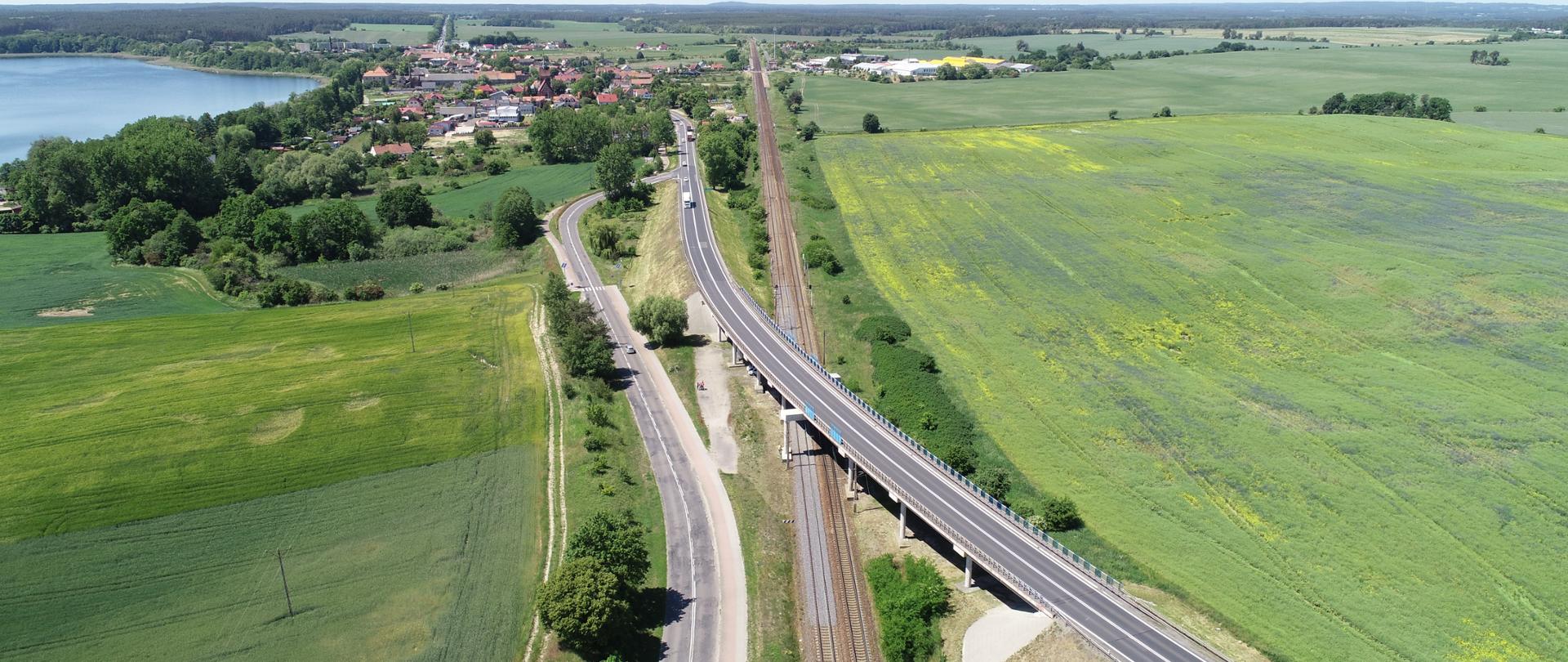 Wiadukt nad linią kolejową w ciągu DK92. Zdjęcie z lotu ptaka krzyżujących się dróg otoczonych polami. W oddali widoczne zabudowania oraz małe jezioro.