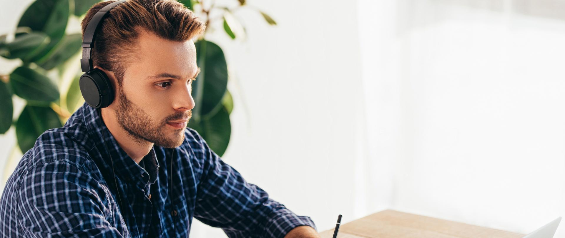 side view of man in headphones taking part in webinar at tabletop with notebook in office