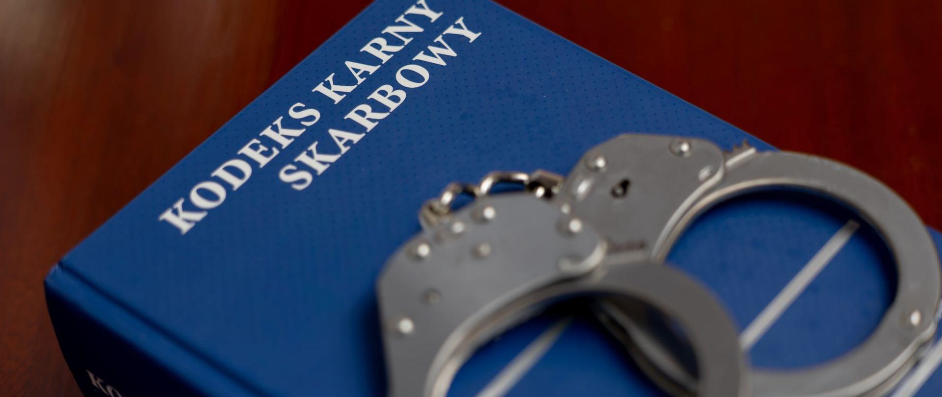 Niebieska książka z napisem „KODEKS KARNY SKARBOWY”. Na książce znajdują się metalowe kajdanki.