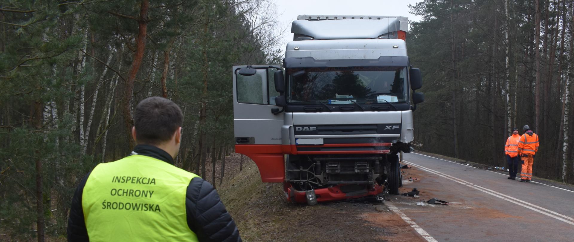 Inspektor Wojewódzkiego Inspektoratu Ochrony Środowiska w Warszawie prowadzi oględziny wypadku samochodu ciężarowego na drodze prowadzącej przez las.