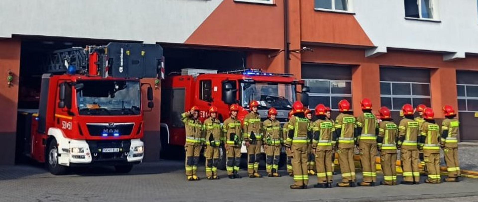 Zdjęcie przedstawia strażaków ustawionych w szyku. W tle samochody pożarnicze. 