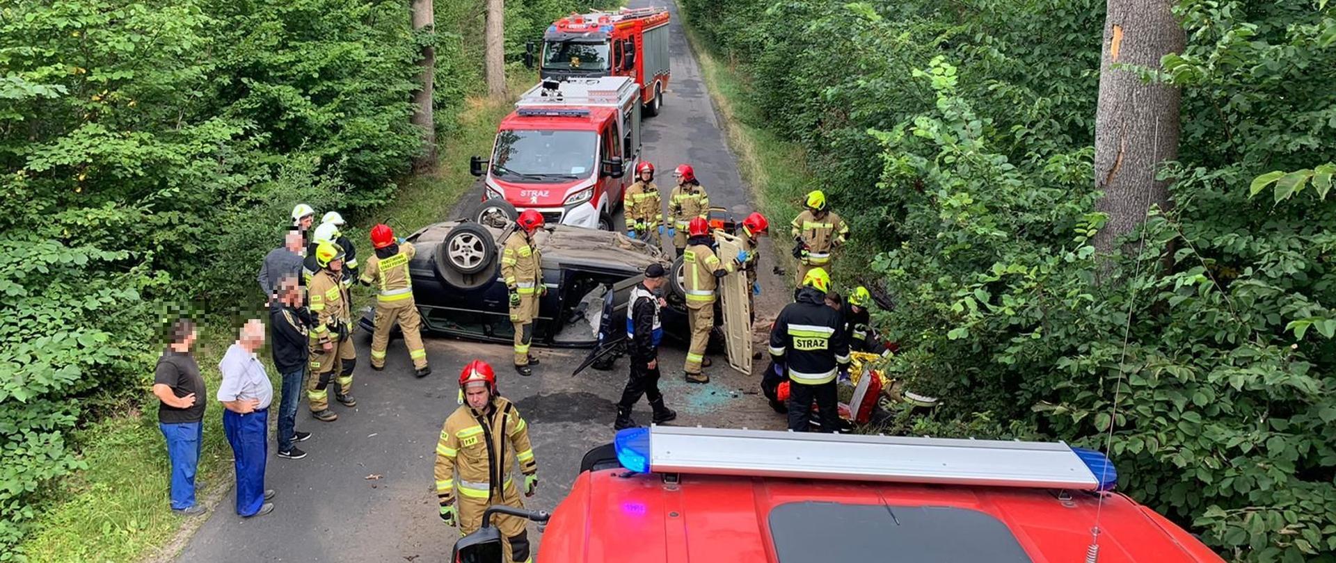 Zdjęcie zrobione z dachu samochodu pożarniczego - strażacy udzielający pomocy medycznej poszkodowanemu oraz prowadzący działania na samochodzie osobowym leżącym na dachu.