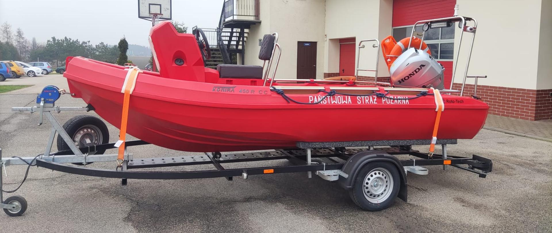 Zdjęcie przedstawia łódź ratowniczą na przyczepce, razem z silnikiem zaburtowym i osprzętem, na tle garażu KP PSP.