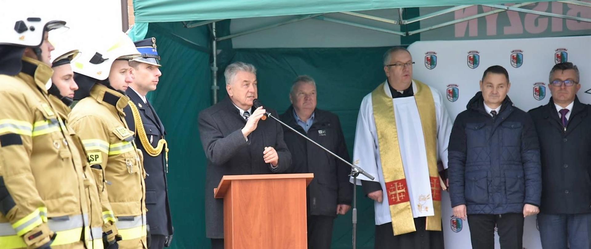 Wicewojewoda przemawia stojąc pod zielonym namiotem. Stoi przy mównicy i trzyma mikrofon lewą dłonią. 