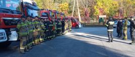 Na zdjęciu strażacy podczas odprawy przed ćwiczeniami taktyczno-bojowymi pk „Skażenie 2022" stojący na zbiórce za nimi czerwone samochody pożarnicze.