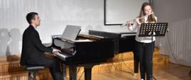 Na zdjęciu wykonanym w auli PSM widnieje uczennica grająca na flecie poprzecznym z akompaniamentem fortepianu. Przy fortepianie nauczyciel Mateusz Gałuszka. Kolorystyka zdjęcia jest biało-czarno-brązowa.
