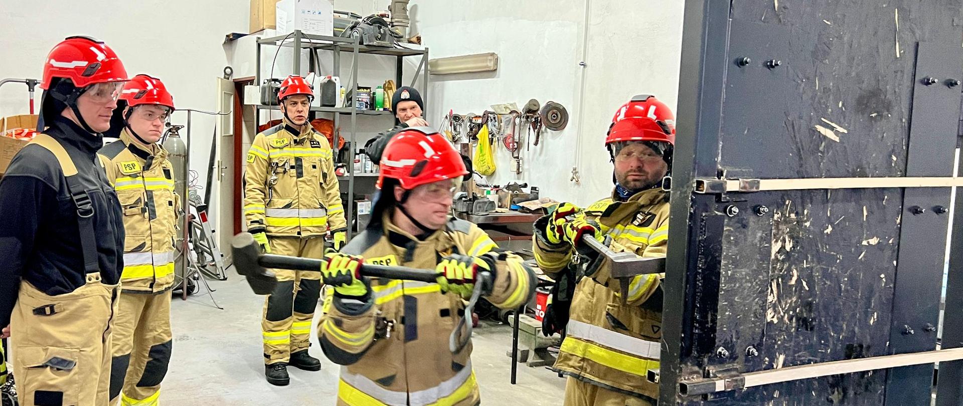 Zmiana I z Jednostki Ratowniczo Gaśniczej ćwiczy siłowe otwieranie drzwi przy pomocy specjalistycznych narzędzi. Strażacy ubrani są w stroje bojowe typu nomex, czerwone hełmy na głowie, na oczach przyłbice. Ćwiczenia odbywają się na garażu Jednostki Ratowniczo Gaśniczej.