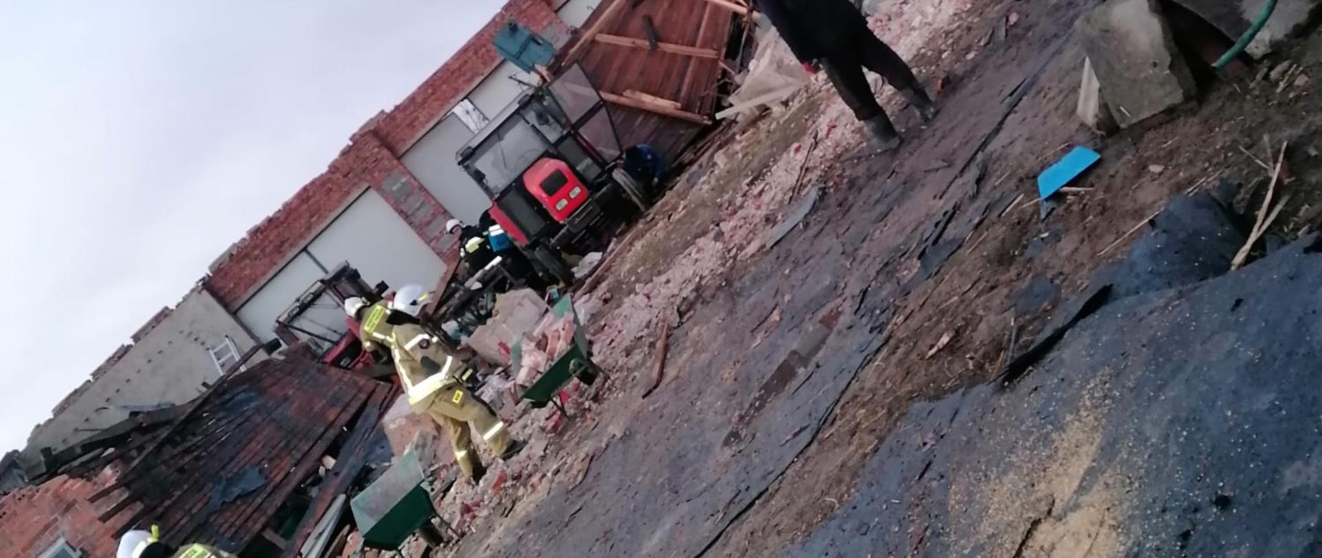 Na zdjęciu widać uszkodzony dach. Strażacy w ubraniach specjalnych - kolor piaskowy przybijają papę i deski. Zachmurzenie duże. Między ścianami uszkodzonego budynku maszyny rolnicze i czerwony traktor. Na pierwszym planie uszkodzone elementy blachy z dachu budynku.