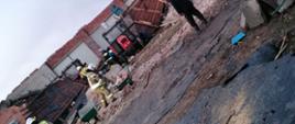 Na zdjęciu widać uszkodzony dach. Strażacy w ubraniach specjalnych - kolor piaskowy przybijają papę i deski. Zachmurzenie duże. Między ścianami uszkodzonego budynku maszyny rolnicze i czerwony traktor. Na pierwszym planie uszkodzone elementy blachy z dachu budynku.