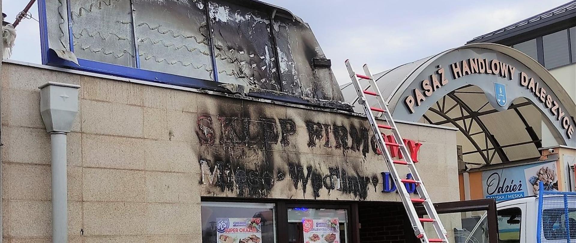 Zdjęcie przedstawia fragment wejścia do sklepu. Nad nim widać spalony, podświetlany kaseton oraz zniszczone litery tworzące nazwę sklepu. Do budynku przystawiona jest drabina, która posłuży do demontażu zniszczonego szyldu.