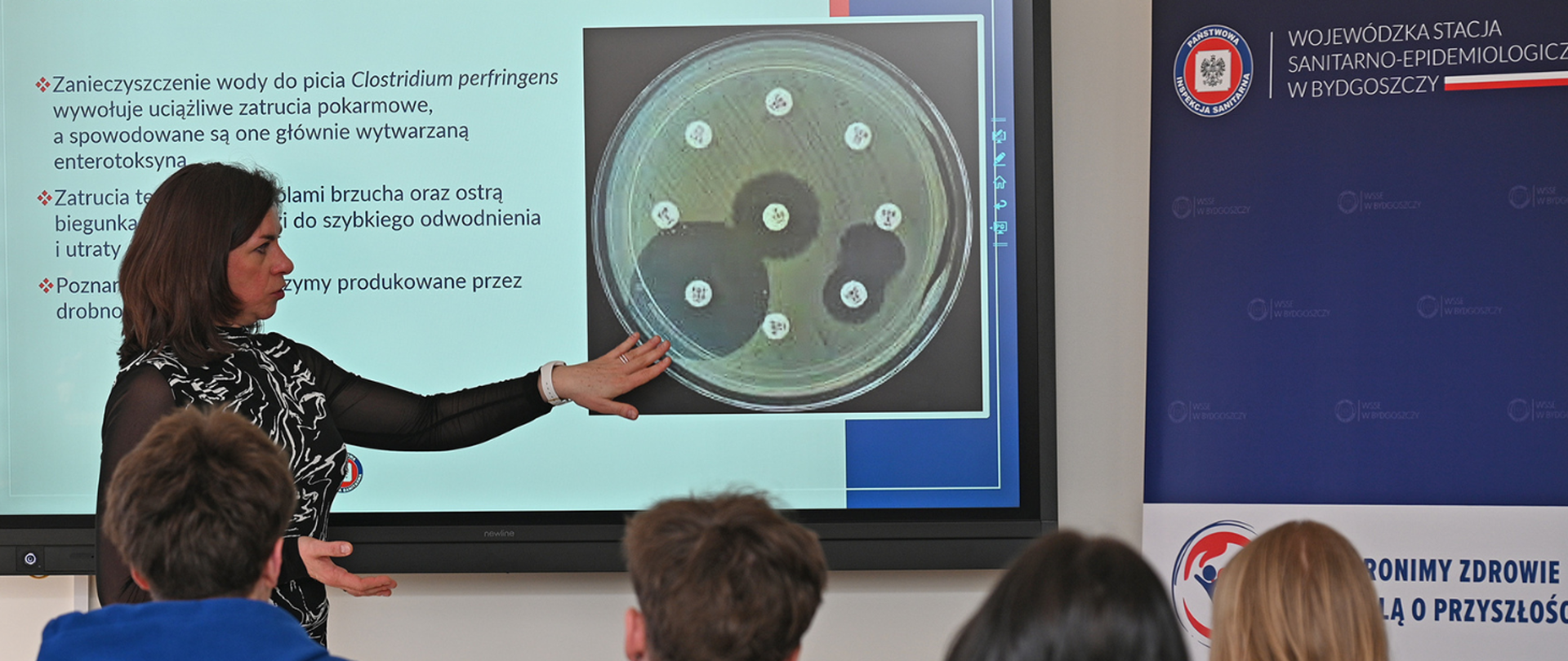 Aneta Gawrysiak-Koszczka prowadzi wykład o mikrobiologicznych badaniach wody dla uczniów, wskazując na ekranie prezentację z informacjami o zanieczyszczeniach wody i zdjęciem próbki laboratoryjnej; obok widoczny baner WSSE w Bydgoszczy.