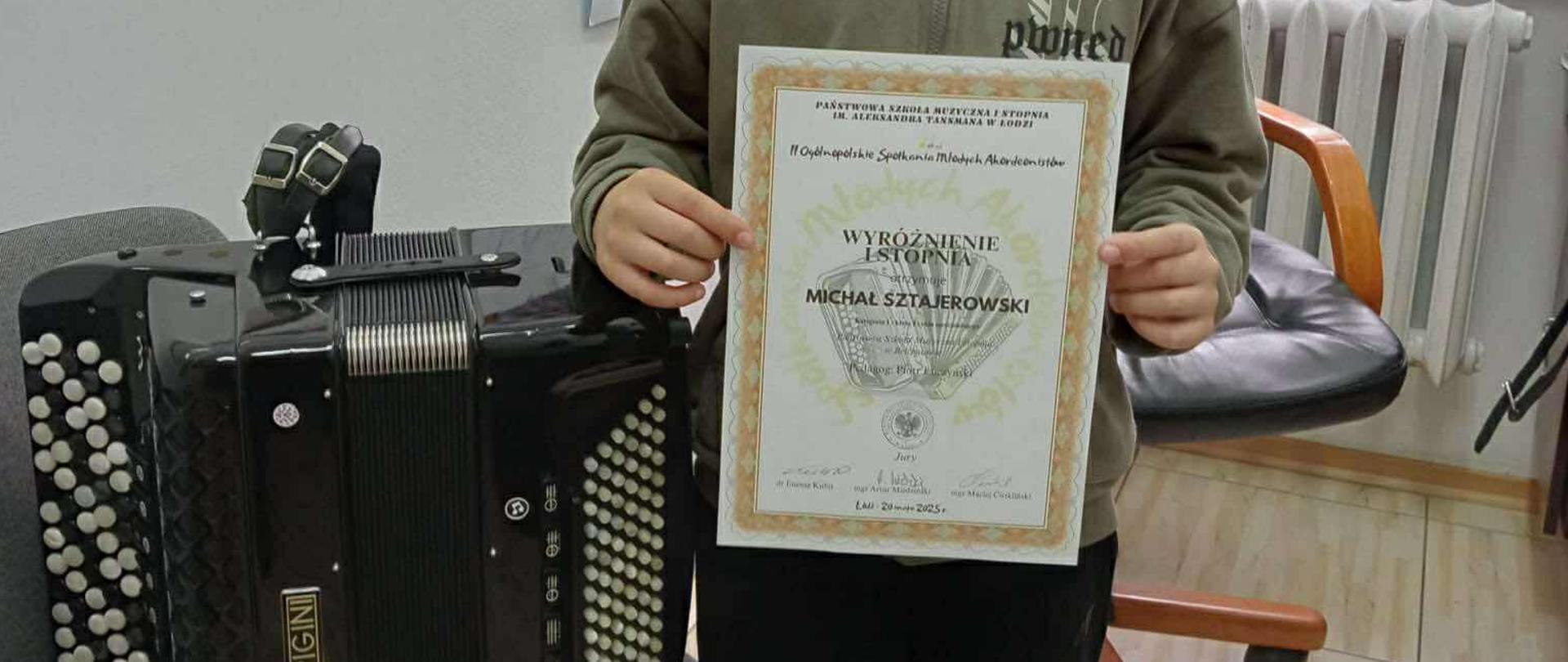 Na zdjęciu znajduje się chłopiec stojący w klasie z dyplomem w rekach. Obok niego na krześle stoi akordeon, a w tle widać na tablicy powieszone rysunki i dyplomy.