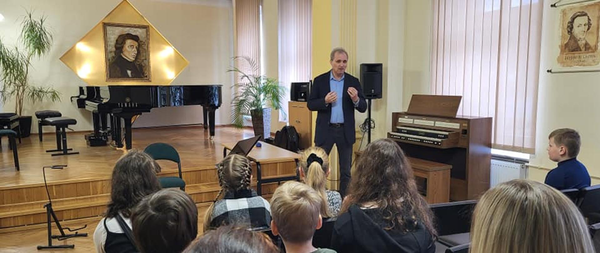 Zdjęcie z warsztatów gitarowych z dr Franciszkiem Wieczorkiem, które odbyły się 4 marca 2023. Zdjęcie wykonane w auli PSM w Kłodzku, na pierwszym planie siedzący uczniowie, którzy słuchają wykład wykładowcy w dalszym planie widoczny fortepian i na ścianie portret F.,Chopina.