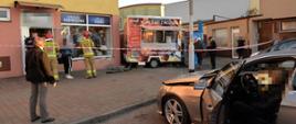 Kolizja z przyczepą gastronomiczną. Parking przed kompleksem handlowo-usługowym na ul. Westerplatte w Rawiczu. Samochód osobowy wjechał na chodnik uszkadzając ścianę i witrynę sklepu oraz przepchnął przyczepę gastronomiczną. Widok na sklep i przyczepę. Przed sklepem strażacy i policjanci oraz cywile. Na parkingu znajduje się auto osobowe biorące udział w kolizji.