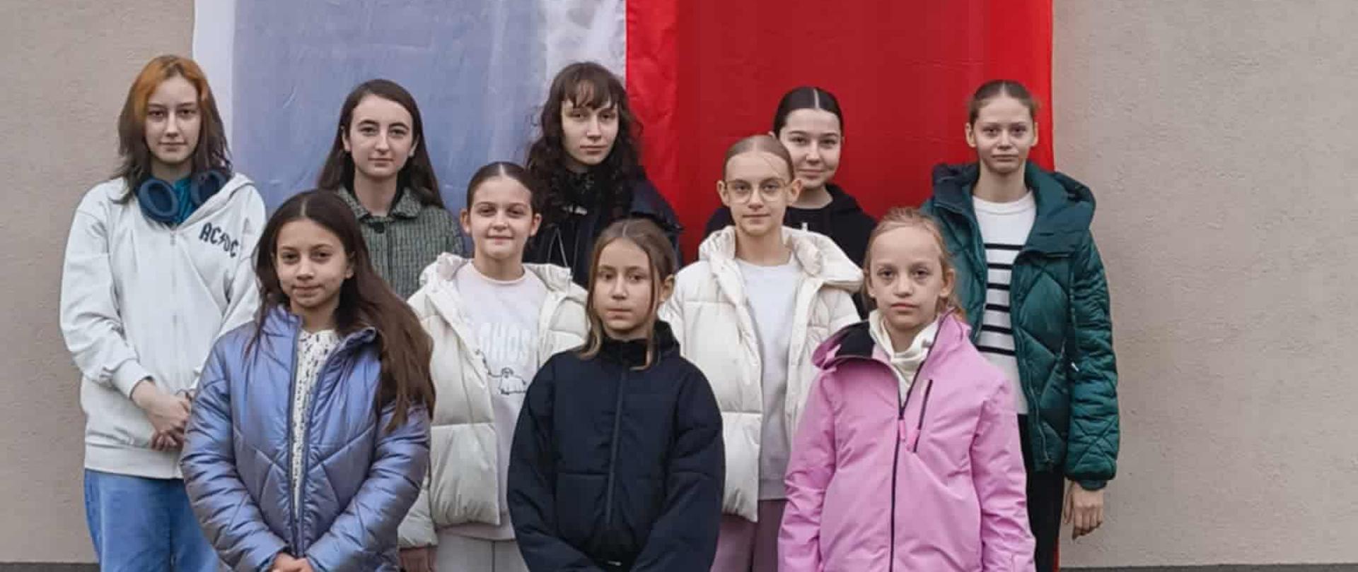 Na zdjęciu grupa dziewcząt stoi zamyślona na tle dużej flagi biało - czerwonej.