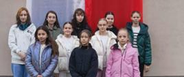 Na zdjęciu grupa dziewcząt stoi zamyślona na tle dużej flagi biało - czerwonej.