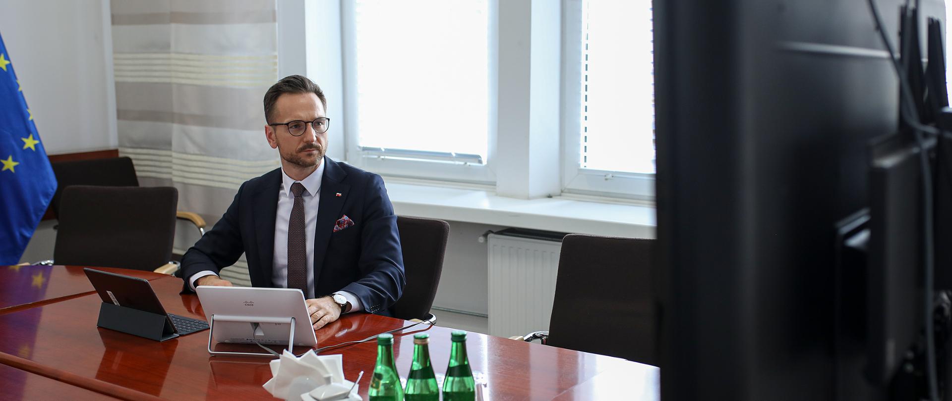 Minister Buda siedzi przy stole patrzy w kierunku telewizora 