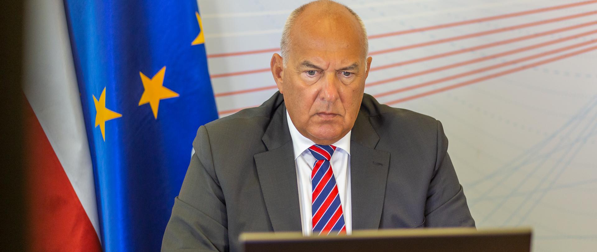 Minister Tadeusz Kościński przed laptopem w gabinecie, w tle flagi Polski i UE 