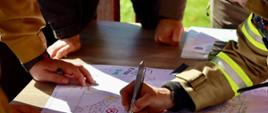 Ujęcie z bliska na stół, na którym rozłożona jest szczegółowa mapa sztabowa oraz dokumenty operacyjne. Dłonie strażaków w mundurach bojowych i służbowych zaznaczają punkty na mapie za pomocą długopisów i markerów, koordynując działania ratownicze. Na planie widoczne są ręczne adnotacje, oznaczenia taktyczne oraz zakreślone obszary.