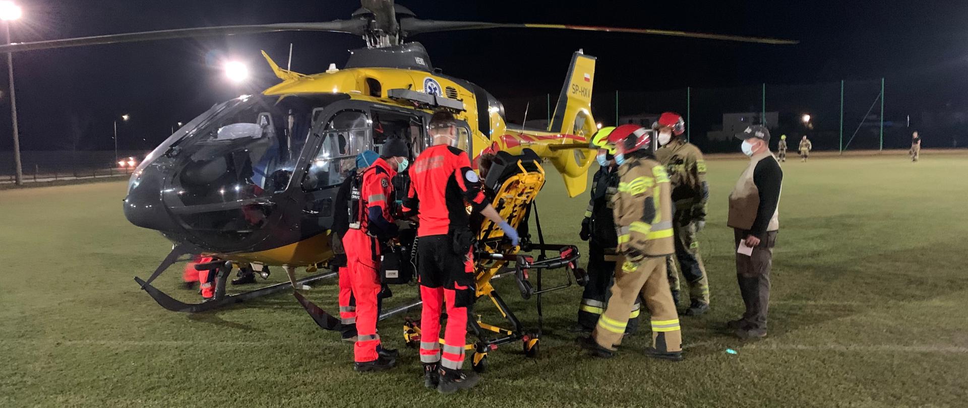 Do śmigłowca LPR przekazywany jest poszkodowany chłopiec leżący na noszach przewoźnych z karetki pogotowia. Obok noszy po lewej stronie stoi dwóch ratowników PRM, po prawej trzech strażaków oraz osoba cywilna. Ratownicy PRM w pomarańczowych uniformach. 2 ratownicy PSP w ubraniach specjalnych koloru piaskowego0 i czerwonych hełmach, trzeci ze strażaków - ratownik OSP w hełmie żółto-zielonym i granatowym ubraniu specjalnym. 