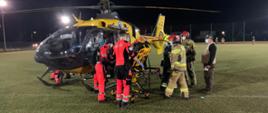 Do śmigłowca LPR przekazywany jest poszkodowany chłopiec leżący na noszach przewoźnych karetki pogotowia. Obok noszy po lewej stronie stoi dwóch ratowników PRM, po prawej trzech strażaków oraz osoba cywilna. Ratownicy PRM w pomarańczowych uniformach. 2 ratownicy PSP w ubraniach specjalnych koloru piaskowego0 i czerwonych hełmach, trzeci ze strażaków - ratownik OSP w hełmie żółto-zielonym i granatowym ubraniu specjalnym. 