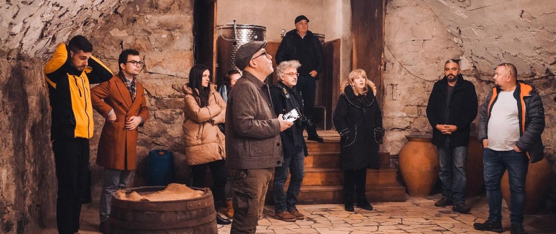 Dziesięcioro, ciepło ubranych osób zwiedza piwnicę z winami.