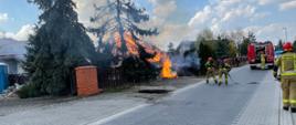 zdjęcie przedstawia ulicę na której stoi samochód strażacki oraz dwóch strażaków w ubraniach specjalnych z aparatami powietrznymi na plecach rozwijają linie i przygotowują się do gaszenia palącej altany . widoczne duże płomienie