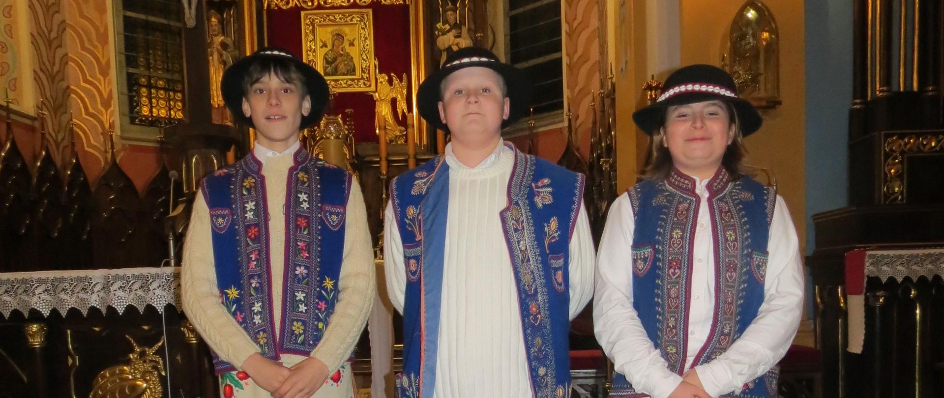 Na zdjęciu widać trzy osoby stojące obok siebie w kościele, ustawione frontem do obiektywu. Znajdują się przed bogato zdobionym ołtarzem w stylu barokowym lub neobarokowym. Ołtarz jest wysoki, złocony, z licznymi ornamentami. W górnej części widoczny jest krucyfiks z postacią Jezusa. W tle są także obrazy religijne, kolumny oraz ciepłe, żółte światło lamp, które nadaje wnętrzu uroczysty, podniosły charakter. Podłoga jest ciemna i lekko błyszcząca. Trzy osoby mają na sobie tradycyjne stroje ludowe: Każda nosi czarny kapelusz z ozdobną, jasną taśmą. Mają białe koszule oraz ciemnoniebieskie haftowane kamizelki z kolorowymi wzorami. Dwie osoby (po lewej i w środku) mają jasne, beżowe spodnie z haftami przy nogawkach. Osoba po prawej ma ciemne spodnie, prawdopodobnie czarne. Postawa wszystkich jest spokojna i wyprostowana, ręce złożone z przodu. Wyraz twarzy neutralny lub lekko uśmiechnięty. Całość sprawia wrażenie uroczystego wydarzenia, być może występu lub ceremonii religijno-kulturalnej.