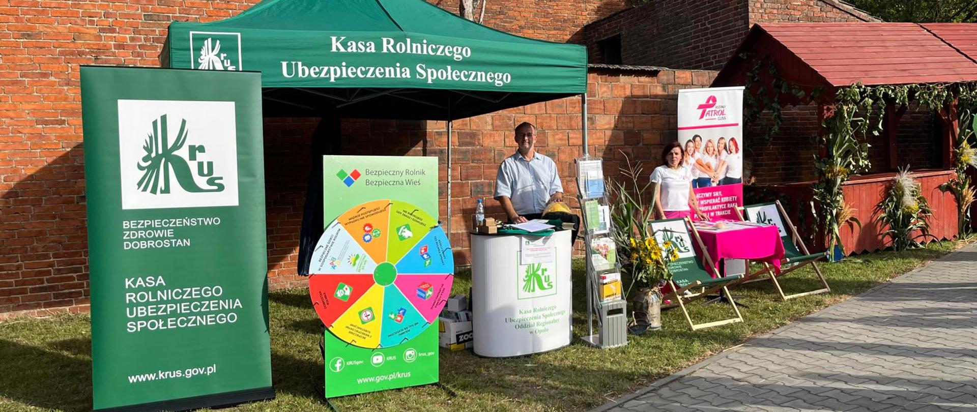 Przy murze z czerwonej cegły pod zielonym namiotem KRUS, za pulpitem z logo KRUS stoi mężczyzna. Obok stoi kartonowa makieta koła fortuny oraz zielony rollup KRUS.
