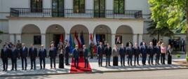 Na czerwonym dywanie stoją premierzy: Donald Tusk i Péter Magyar. Po lewej i prawej stronie premierów stoją członkowie rządu Polski i Węgier. W tle 9 flag (3 flagi Polski, 3 Węgier i 3 UE). Przy flagach stoi 6 żołnierzy (dwie trójki). W tle fasada budynku z tarasem oraz inne osoby. Po prawej stronie drzewa.
