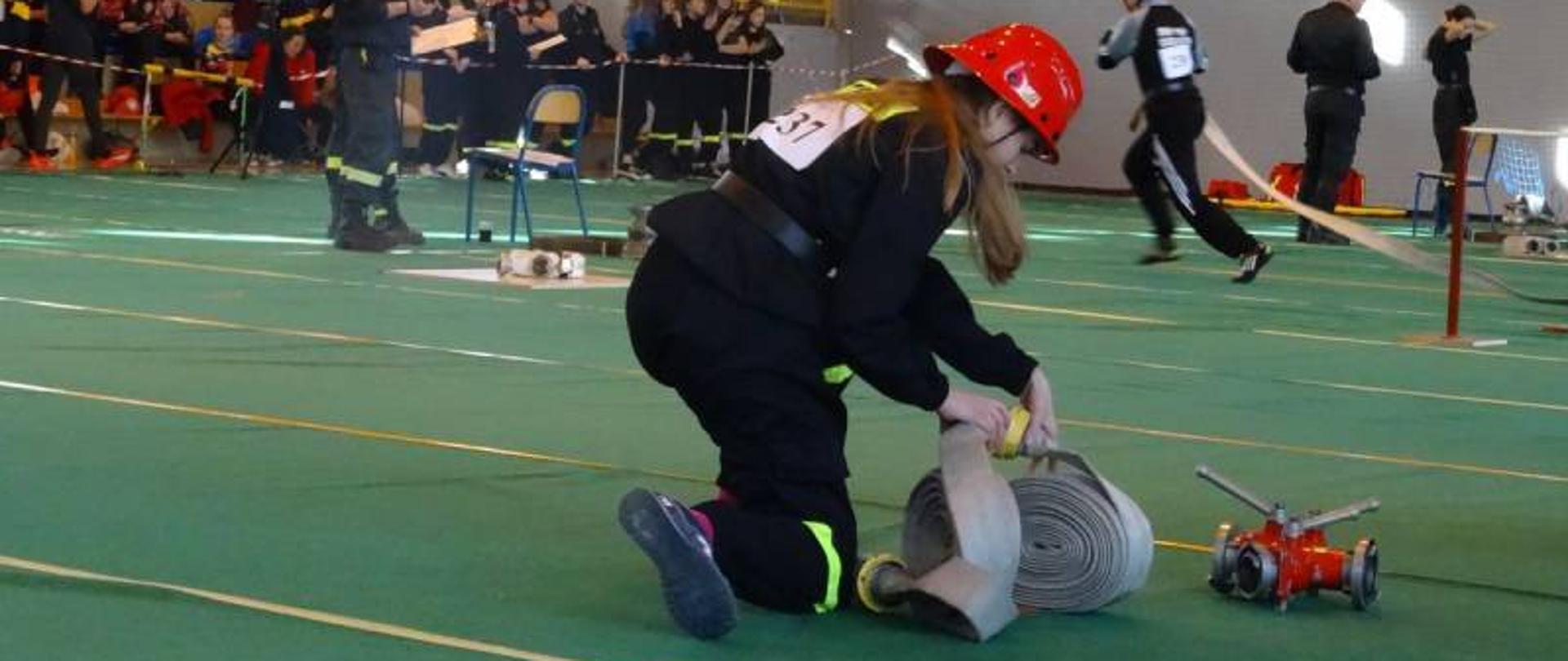 Strażak MDP łączy węże. W tle inny strażacy MDP wykonują zadania.Na trybunach siedzą młodzieżowcy.