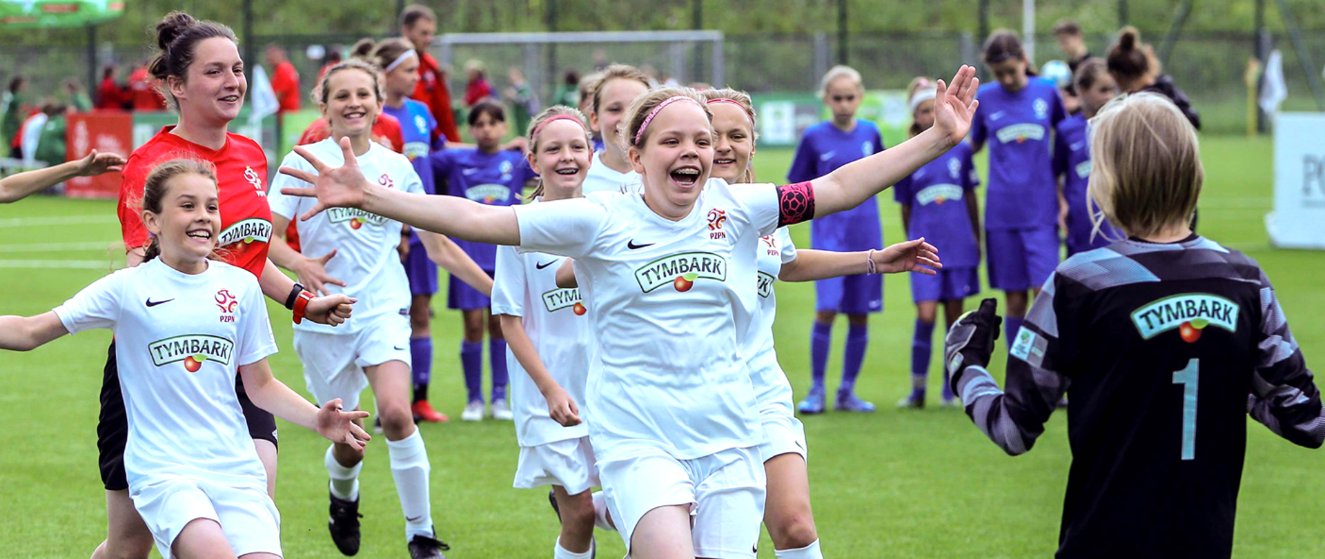 Boisko piłkarskie. Radosne dzieci w koszulkach Tymbarku biegną w kierunku stojącego tyłem do aparatu bramkarza. W tle stoją inni zawodnicy w fioletowych strojach.
