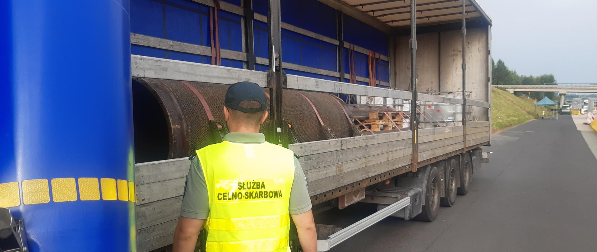 Osoba w kamizelce z napisem „Służba Celno-Skarbowa” stoi obok otwartej ciężarówki. Na naczepie znajdują się duże metalowe elementy transportowane i zabezpieczone pasami mocującymi.