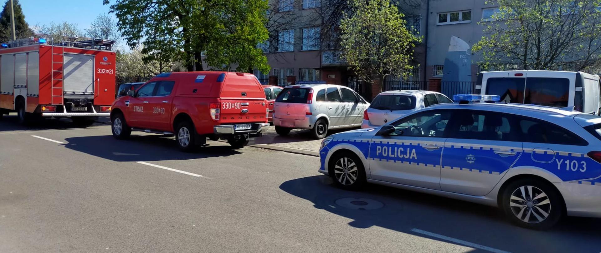 Zdjęcie wykonane przed jedną za szkół na terenie miasta Radomia. Przed szkołą ustawione są dwa pojazdy należące do Państwowej Straży Pożarnej oraz jeden do Policji. Pierwszy pojazd jest to samochód ciężarowy przeznaczony do działań ratowniczo - gaśniczych. Pojazd w kolorze czerwonym ze srebrnymi elementami zabudowy. Drugi to pojazd terenowy typu pick-up. Pojazd również jest w kolorze czerwonym. Za nimi ustawiony jest radiowóz Policji. Samochód osobowy typu kombi w kolorze srebrno - niebieskim.

