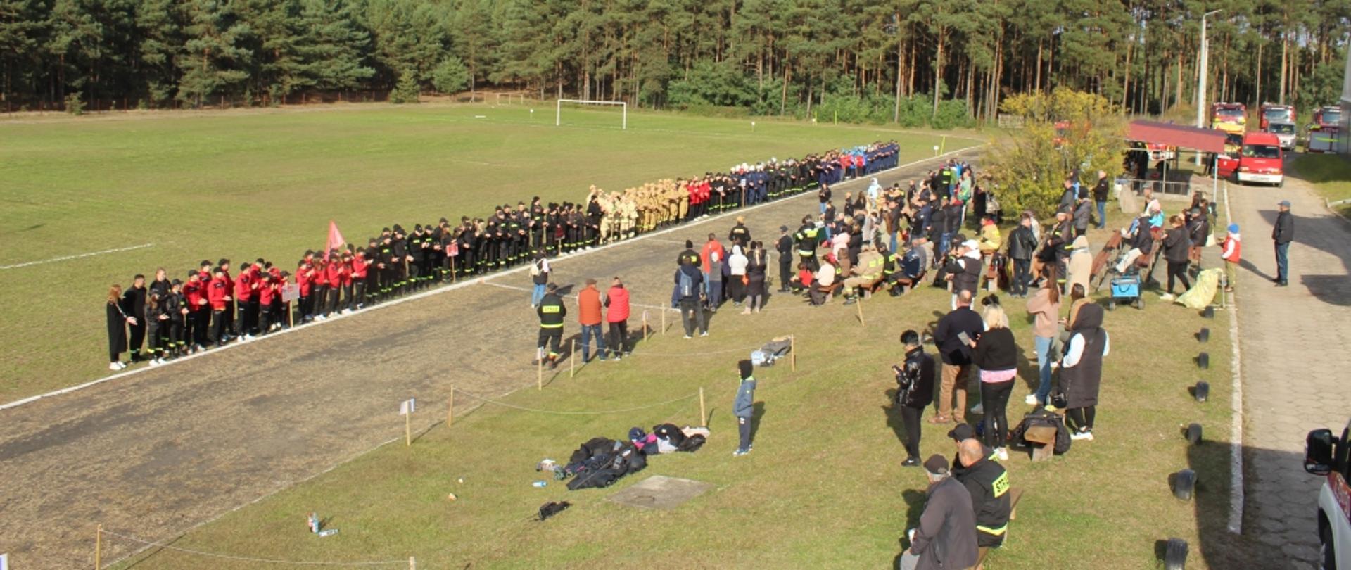 Szeroka perspektywa ukazuje uroczyste rozpoczęcie wydarzenia. Na pierwszym planie, wzdłuż bieżni, stoją w szyku dwie duże grupy uczestników, ubranych głównie w mundury w kolorach czerwonym i czarnym (lub ciemnym), oraz beżowym. Po prawej stronie widać zgromadzoną publiczność i część infrastruktury, w tym wozy strażackie. W tle rozciąga się duża, trawiasta murawa i las.