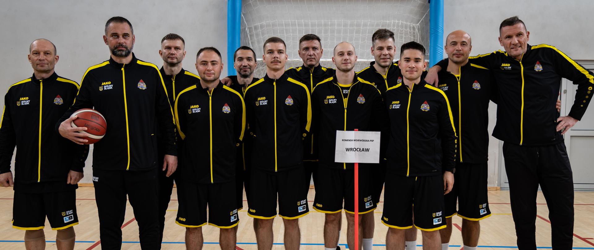 Grupa dwunastu mężczyzn w czarno-żółtych dresach sportowych pozuje do zdjęcia na hali sportowej, stojąc za tabliczką z napisem „WROCLAW”