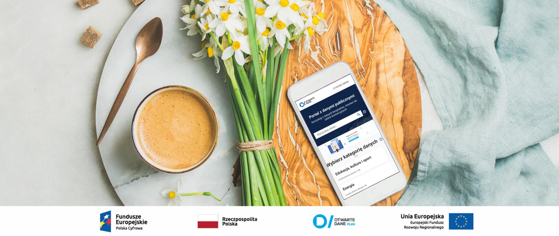 Zdjęcie z góry - stolik, na którym stoi filiżanka kawy, złota łyżeczka, bukiet żonkili oraz telefon komórkowy, na ekranie którego widoczna jest strona dane.gov.pl. 